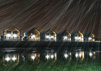 Cabins lit up at night with reflections in the water, seen through a blurred foreground.
